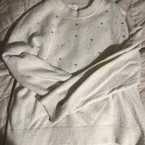 Vit stickad tröja med pärlor H&M - Superfin vit stickad tröja från H&M med pärldetaljer framtill. Tröjan har rund halsringning, långa ärmar och ribbade muddar vid ärmslut och nederkant. Perfekt för dig som gillar en clean look med lite extra bling.