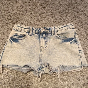 Ljusblå jeansshorts med fransig kant - Snygga ljusblå jeansshorts med slitna, fransiga kanter och klassisk femficksmodell. Shortsen har bälteshällor och en cool tvättad look som ger en avslappnad vibe. Perfekta för varma dagar när du vill ha en chill och trendig stil.