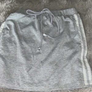 Grå sportig kortkjol med snörning XS - Grå kortkjol i mjuk bomullsmix med elastisk midja och snörning. Snygga vita ränder längs sidorna ger en sportig vibe. Perfekt för en chill och avslappnad stil. Passar dig som gillar bekväma och trendiga plagg.