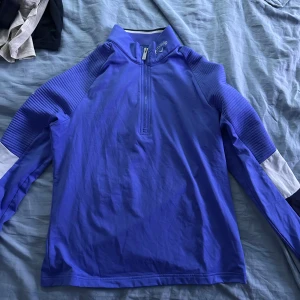 Blå långärmad tröja från Callaway - Sportig blå långärmad tröja från Callaway med ribbade detaljer på ärmarna och vit panel på underarmen. Tröjan har en hög krage och half zip framtill, perfekt för golf eller andra utomhusaktiviteter.