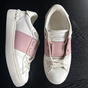 Valentino Garavani sneakers vit/rosa 36,5 - Snygga Valentino Garavani sneakers i vitt skinn med breda ljusrosa detaljer över vristen. Klassisk rund tå, vita snören och coola nitar på hälen. Perfekta för dig som vill ha en lyxig och trendig look med en touch av färg.