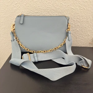 Ljusblå axelväska med guldkedja - Snygg ljusblå axelväska i skinn med bred justerbar rem och guldfärgad kedjedetalj. Väskan har dragkedja upptill och två fack inuti. Väskan är ifrån Mango och endast använd 2 gånger. Pris går att diskutera!