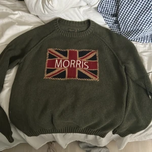 Olivgrön stickad tröja från Morris - Snygg olivgrön stickad tröja från Morris med Union Jack-flagga och broderad logga på bröstet. Tröjan har rund halsringning och ribbade muddar vid ärmslut och nederkant. Perfekt för dig som gillar klassisk brittisk stil och är en 100% riktig tröja