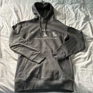 Mörkgrå hoodie storlek M - Snygg mörkgrå hoodie med vit text på bröstet och klassisk huva med snörning. Tröjan har långa ärmar och ribbade muddar. Perfekt för chill dagar eller när du vill ha en enkel och stilren look.