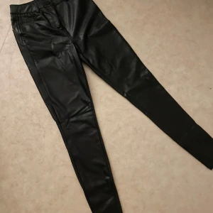 Svarta skinnleggings med tight passform - Snygga svarta leggings i skinnimitation med hög midja och tight passform. Byxorna har dragkedja och knapp framtill samt smala ben som sitter som en andra hud. Från new yorker