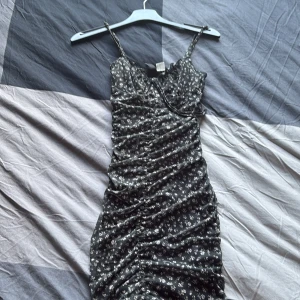 Svart blommig kortklänning med smala axelband - Supersöt svart kortklänning med små vita blommor och smala axelband. Klänningen har rynkade detaljer längs sidorna som ger en snygg passform och en feminin look. Perfekt för dig som gillar en trendig och somrig stil!