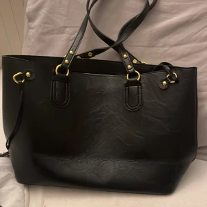 Svart axelväska med gulddetaljer - Stilren svart axelväska i skinnimitation med snygga guldfärgade metalldetaljer och dubbla axelremmar. Väskan har en rymlig insida och klassisk form, perfekt för att bära med sig allt man behöver. Enkel men ändå elegant design som passar till många outfits.