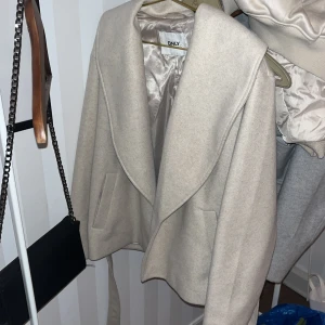 Beige kappa från ONLY med bälte - Snygg beige kappa från ONLY med bred krage och bälte i midjan. Jackan har en stilren design med två fickor framtill och är helfodrad. Perfekt för dig som gillar en clean och trendig look.