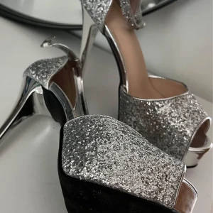 Glittriga silverfärgade platåpumps - Snygga pumps med hög klack och platåsula i silver. Skorna är täckta av glittrigt material och har öppen tå samt smal ankelrem. Perfekta för dig som vill sticka ut och glänsa. Klacken är riktigt hög och ger extra attityd till outfiten.
