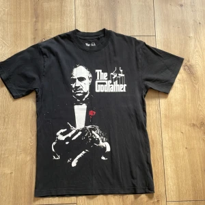 Svart The Godfather t-shirt loose fit - Svart t-shirt med stort The Godfather-tryck fram och bak. Loose fit-modell med klassisk rund hals och korta ärmar. Ikoniskt filmtryck i vitt och rött som sticker ut. Perfekt som gillar Grisch och Old style stilarna😃