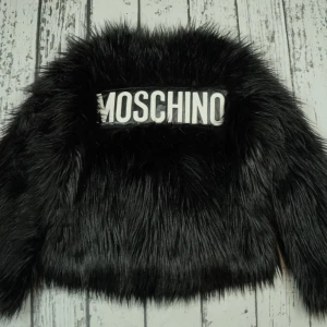 Svart pälsjacka från Moschino x H&M - Säljer en svart pälsjacka från Moschino x H&M med stor logga på ryggen. Jackan har lång, fluffig fuskpäls och en oversized vibe. Perfekt statement-plagg för dig som vill sticka ut och älskar streetstyle. Kedjedetalj i nacken för extra edge.