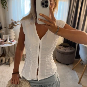 Vit blus med knappar från Weekday - Säljer en vit blus från Weekday i storlek 38. Toppen har en figurnära passform med snygg rynkdetalj längs sidorna och knappar framtill. Ärmlös design med v-ringning, perfekt till jeans eller kjol för en clean look. Passar mig som har 34-36