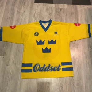 Svenska Tre Kronor hockeytröja gul/blå - Gul hockeytröja med blå detaljer och Tre Kronor-tryck på bröstet. V-ringad hals, breda ärmar och stora loggor från Oddset och Coca-Cola. Perfekt för dig som vill sticka ut på match eller sportevent. denna modellen bars under 90-/ tidigt 2000tal