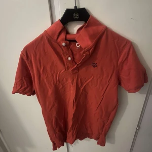 Röd pikétröja från C&A - Snygg röd pikétröja från C&A med klassisk krage och två knappar framtill. Tröjan har korta ärmar och en liten broderad logga på bröstet. Perfekt för dig som gillar en enkel och stilren look.