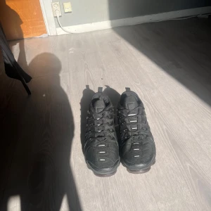 Svarta Nike Air Vapormax sneakers - Säljer ett par helsvarta Nike Air Vapormax sneakers med synlig luftbubbelsula och coola detaljer på ovandelen. Skorna har snörning, rund tå och är tillverkade i syntet och textil. Perfekta för dig som vill ha en sportig och modern look.