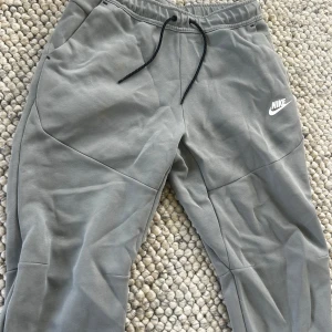 Gråa Nike-Tech byxor oanvänd medium  - Snygga grå mjukisbyxor från Nike med svart snörning i midjan och vit logga på låret. Byxorna har en relaxed fit och är perfekta för chill eller träning. Tillverkade i mjukt bomullsmaterial för extra komfort.