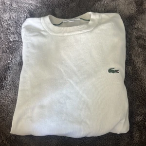 Vit tröja Lacoste  - Säljer en stilren vit tröja från Lacoste med klassisk rund hals och den ikoniska gröna krokodilloggan broderad på bröstet. Tröjan är i bomull och har en normal passform, perfekt för en clean stil.