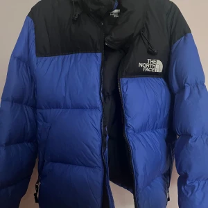 Blå dunjacka från The North Face (Nuptse) - Säljer en blå och svart dunjacka från The North Face. Jackan har en klassisk pufferstil med hög krage, dragkedja och logga på bröstet och ryggen. Perfekt för kalla dagar och riktigt snygg till streetwear-looken.