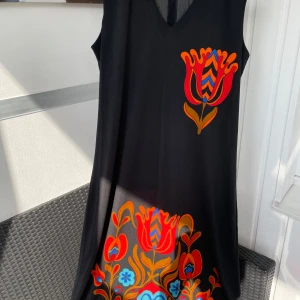 Svart klänning med färgglada blommor Holly & Whyte - Svart ärmlös kortklänning från Holly & Whyte by Lindex med färgglada blommiga tryck i rött, orange och blått. Klänningen har v-ringning och ett snyggt blommönster både fram och bak. Perfekt för dig som gillar retrovibbar och färg.