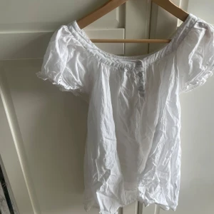 Vit blus med puffärm från Gina Tricot - Supersöt vit blus från Gina Tricot med puffiga korta ärmar och knappar framtill. Den har en bred, rundad halsringning och är gjord i tunn bomull som känns luftig och lätt. Perfekt för en fräsch och enkel look.