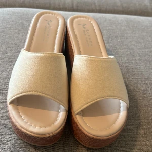 Beige plattform sandaler från Aoerda - Snygga sandaler med beige ovandel och bred rem, öppen tå och plattformsula i brunt med mönstrad struktur. Perfekta för sommaren och ger extra höjd med sin chunky plattform. Ovandelen är i syntetmaterial och sulan har ett coolt flätat utseende.