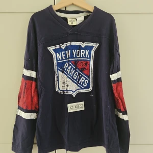 New York Rangers tröja från CCM - Snygg långärmad New York Rangers tröja från CCM i mörkblått med stora tryck på bröstet och armarna i rött, vitt och blått. Tröjan har V-ringning och är gjord i 100% bomull. Perfekt för dig som älskar hockey och vill sticka ut med sportig stil.