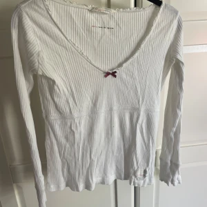 Odd Molly topp - Supersöt vit ribbad långärmad topp från Odd Molly med v-ringning, spetskant och en liten vinröd rosett framtill. Broderad detalj på ärmen med blommor och text.