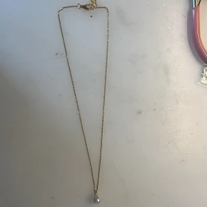 Guldhalsband med vit pärla - Rostfritt stål. Stilrent halsband i guldtonad kedja med en droppformad vit pärla som hänge. Perfekt för dig som gillar klassiska smycken med en modern twist. Kedjan är tunn och elegant, och pärlan ger en lyxig känsla till din look. Handgjord