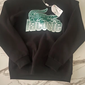 Svart sweatshirt från Lacoste med logga - Svart sweatshirt från Lacoste i bomull med stor grön och vit Lacoste-logga framtill. Klassisk rund halsringning och långa ärmar med ribbade muddar. Perfekt för dig som gillar streetstyle och vill ha en clean look med tydlig branding.