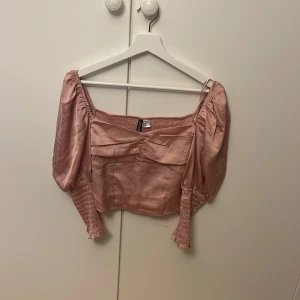 Rosa satinblus från H&M Divided - Supersöt ljusrosa blus i satin från H&M Divided. Croppad modell med puffiga långärmar, smockade muddar och smockad rygg. Snygg omlottkänsla framtill och lätt glansigt material som ger en lyxig vibe. Perfekt för dig som gillar romantisk och trendig stil.