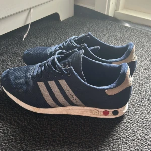 Adidas LA Trainer blå sneakers - Snygga Adidas LA Trainer sneakers i marinblått med klassiska tre vita ränder på sidan och detaljer i silver. Ovandel i mesh för bra andningsförmåga, snörning och vit sula med färgade pluggar. Perfekta för dig som gillar retrostil och sportig vibe.