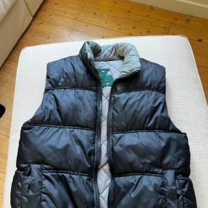 Svart dunväst från Authentique - Snygg svart dunväst från Authentique med quiltad insida i grått och broderad logga på kragen. Västen har dragkedja framtill och en klassisk puffer-look som ger extra volym. Perfekt att slänga över en hoodie eller tröja när det är kyligt ute.