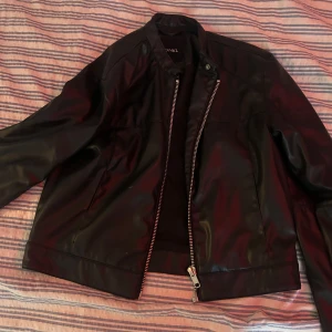 Svart skinnjacka från MONKI - Snygg svart skinnjacka från MONKI med raka linjer och silverfärgad dragkedja. Jackan har en klassisk krage med tryckknapp och två sidfickor. Perfekt för dig som vill ha en stilren och edgy look. (AI) 