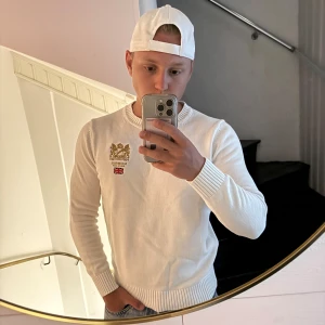 Vit stickad tröja från Morris - Säljer en stilren vit stickad tröja från Morris med broderad gulddetalj och brittisk flagga på bröstet. Tröjan har rund hals, ribbade muddar och är långärmad. Perfekt för dig som gillar klassisk och clean stil.