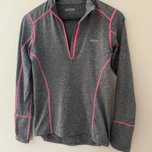 Grå tränings-tröja OTP XS med rosa detaljer - Sportig grå långärmad tröja från OTP i storlek XS. Snygga rosa sömmar och dragkedja framtill (half zip). Perfekt för träning eller löpning, med liten ficka bak och reflekterande detalj. Mjuk och stretchig syntetblandning.