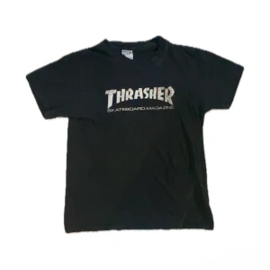 Svart Thrasher t-shirt i bomull - Svart t-shirt med klassiskt Thrasher Skateboard Magazine-tryck på bröstet. Skön passform och tillverkad i 100% bomull, perfekt för dig som gillar streetwear och skatekultur. Enkel och stilren design med korta ärmar.
