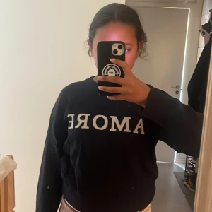 Svart tröja med AMORE från Gina Tricot - Svart långärmad tröja från Gina Tricot med vit texten 'AMORE' framtill. Klassisk rund halsringning och relaxed fit, perfekt för en chill och stilren look. Mjuk och skön, passar till jeans eller mjukisbyxor.