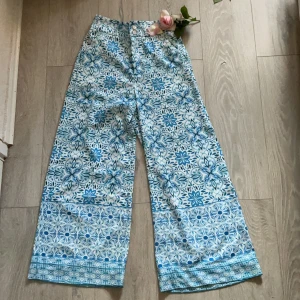 Blåvita vida byxor med blommönster - Säljer ett par riktigt snygga vida byxor med blått och vitt blommönster. Byxorna har hög midja, resår baktill och knäppning framtill med två knappar. Materialet känns som bomull, perfekt för vår och sommar. Unik och fräsch look som sticker ut!
