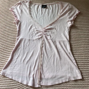 Ljusrosa topp med spets från Gina Tricot - Supersöt ljusrosa topp från Gina Tricot med v-ringning, spetskant och rynkdetalj framtill. Korta ärmar med vågig kant och en feminin passform. Öppen i mitten. Perfekt att styla med jeans eller kjol för en söt look. Använd några fåtal gånger men ser som ny ut❤️