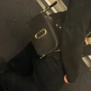 Svart axelväska Marc Jacobs - Säljer en svart axelväska från Marc Jacobs med guldiga metalldetaljer och logga framtill. Väskan har en bred axelrem med text och dragkedja upptill. Perfekt storlek för det viktigaste och riktigt snygg till alla outfits. Priset vid snabb äffär kan gå ner.