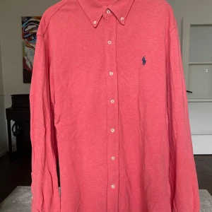 Rosa skjorta från Ralph Lauren - Snygg rosa skjorta från Ralph Lauren i lätt och luftig bomullskvalitet. Klassisk button-down krage, långa ärmar och knappar framtill. Ikoniska blå loggan broderad på bröstet. Perfekt för dig som gillar stilrena och tidlösa plagg.