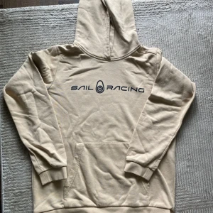Beige hoodie från Sail Racing - Snygg beige hoodie från Sail Racing med svart logga framtill. Klassisk känguruficka och huva, perfekt för chill dagar. Mjuk insida och ribbade muddar vid ärmslut och nederkant. Passar dig som gillar enkel och stilren streetwear.