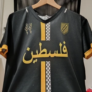 FC Palestina svart/gul fotbollströja - Svart fotbollströja med gula detaljer och tryck från FC Palestina. Tröjan har korta ärmar, V-ringning och unika grafiska tryck på både fram- och baksida, inklusive arabiska bokstäver och kartmotiv. Tillverkad i lätt och ventilerande syntetmaterial.