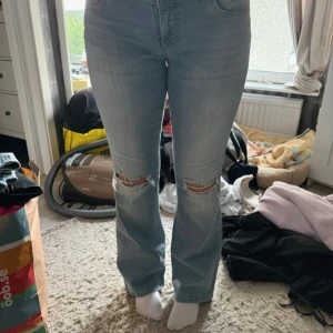 Ljusblå bootcut jeans med slitningar - Säljer ett par ljusblå jeans med bootcut-modell och snygga slitningar över knäna. Jeansen har låg midja  och klassisk femficksdesign. Perfekta för dig som gillar en avslappnad men trendig look.