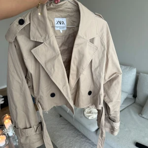 Beige croppad trenchcoat från Zara - Snygg croppad trenchcoat från Zara i beige bomull. Jackan har klassisk krage, knappar framtill och bälte i midjan. Rå kant nertill för en trendig look. 💛