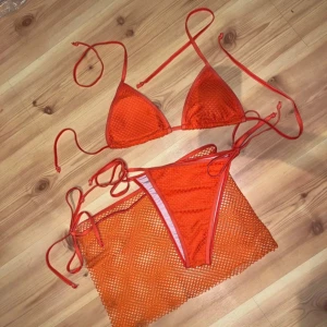 Orange bikini med kjol och snörning - Snygg orange bikini med trekants-bh och knyt i sidorna. Matchande nätkjol ingår för extra sommarvibe. Bikinin har ett coolt meshmönster och tunna band för en trendig look. Perfekt för stranden eller poolhänget!
