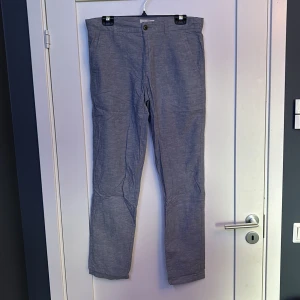 Blå tapered byxor från Jack & Jones - Snygga blå byxor från Jack & Jones i tapered modell. Byxorna har klassisk passform med raka ben, bälteshällor och knäppning fram. Tillverkade i ett lätt och svalt material som passar perfekt till varmare dagar. Fickor både fram och bak med knappar bak. I mycket bra skick men lite skrynkliga.