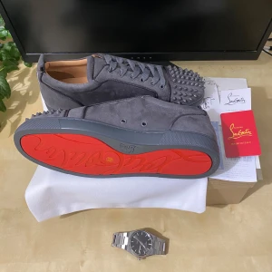 Gråa Christian Louboutin sneakers med nitar - Snygga grå sneakers från Christian Louboutin i mocka med ikoniska röda sulor och coola nitar över tån. Skorna har snörning och en platt sula, perfekt för dig som vill sticka ut med en edgy och lyxig stil.