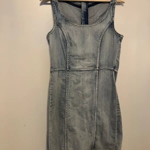 Jeansklänning från Lindex denim  - Snygg ärmlös jeansklänning från Lindex i ljusblå denim. Klänningen har figurnära passform, markerade sömmar och en lång dragkedja bak. Perfekt för dig som gillar en enkel men cool stil.