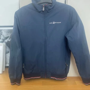 Marinblå vindjacka från Sail Racing - Snygg marinblå vindjacka från Sail Racing med dragkedja framtill och logga på bröstet. Jackan har ribbade muddar med röd och vit detalj, hög krage och två fickor med dragkedja. Perfekt för dig som gillar sportig och stilren look.